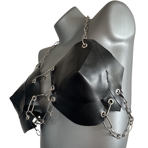 Puede incluir: Sujetador de látex negro con detalles de cadena plateada. El sujetador presenta un diseño geométrico con copas angulares y correas de cadena. Las cadenas conectan las copas y envuelven los hombros y el torso.