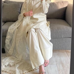Puede incluir: Conjunto de dos piezas color crema con pantalón de pernera ancha y top de manga larga. El conjunto incluye una bufanda transparente bordada. El top tiene mangas abullonadas. El conjunto se lleva con sandalias plateadas.