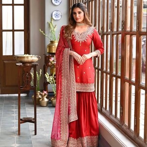 Puede incluir: Un conjunto de salwar kameez rojo bordado. El conjunto incluye una túnica de manga larga, pantalones de pernera ancha y una bufanda a juego. La túnica y los pantalones presentan intrincados bordados dorados a lo largo del escote, los puños y el dobladillo. La bufanda tiene un estampado rosa y dorado.