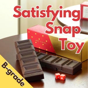 May include: A brown, rectangular snap toy resembling a chocolate bar, with the text "PAKITE" on the bottom. The image also shows two red dice and two gift boxes, one red with gold hearts and the other gold.