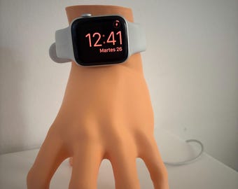 Puede incluir: Un smartwatch gris claro con una pantalla negra que muestra la hora 12:41 y la fecha. El reloj está sobre un soporte en forma de mano de color melocotón con uñas pintadas de blanco. Se ve un cable de carga.