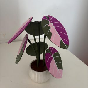 Op de afbeelding: Een decoratieve kunstplant met grote, kleurrijke bladeren in roze, paarse en groene tinten. De bladeren zijn bevestigd aan donkergroene stelen en staan in een witte, geribbelde keramische pot. De plant is een miniatuurversie van een tropische plant.