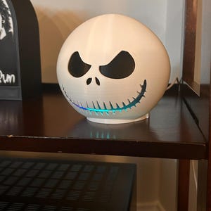 May include: A white, spherical decorative lamp featuring the face of Jack Skellington from The Nightmare Before Christmas. The lamp has black eye and mouth cutouts, and a blue and green light emanates from the bottom. The lamp is sitting on a dark brown shelf.