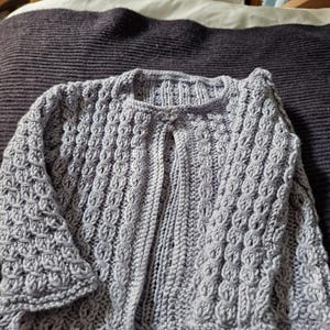 Puede incluir: Un cárdigan tejido a mano de color lila claro con un diseño texturizado. El suéter abierto tiene cuello redondo y mangas acampanadas. El cárdigan está hecho de un hilo suave, posiblemente lana. La prenda se muestra sobre una manta morada oscura.