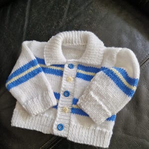 May include: A white knitted baby cardigan with blue and yellow stripes on the sleeves and chest. The cardigan has a collared neckline and is fastened with blue and cream buttons. The garment is made of soft yarn.