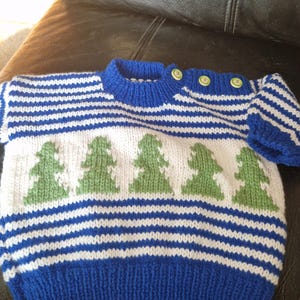 Pode incluir: Um suéter de malha azul, branco e verde, com gola redonda e três botões no ombro. O design inclui listras horizontais azuis e brancas e uma fileira de motivos de árvores verdes. As mangas são curtas.