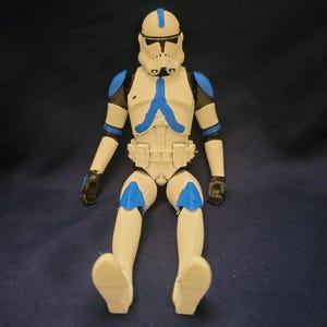 May include: A white and blue action figure of a clone trooper from a science fiction franchise. The figure is in a seated position with its legs extended. The trooper's armor features blue accents on the shoulders, helmet, and other areas. The figure has a detailed helmet and belt.