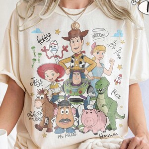 May include: An ivory t-shirt featuring a colorful graphic print of Toy Story characters including Woody, Buzz Lightyear, Jessie, Bo Peep, Forky, Bullseye, Mr. Potato Head, Rex, and Hamm, with their names written in a cursive script font.