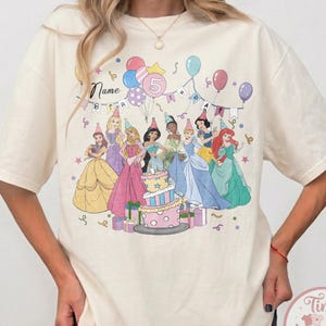 May include: Ivory-colored t-shirt featuring a colorful graphic of Disney princesses around a birthday cake. The design includes balloons, party hats, and the number 5. The word "Name" is also visible on the shirt.