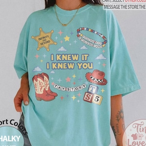 May include: A light mint green t-shirt featuring a graphic print with a yellow sheriff star, a beaded bracelet, a red cowboy boot, and alphabet blocks. The text reads: "INFINITY AND BEYOND", "YOU'VE GOT A FRIEND IN ME", "I KNEW IT I KNEW YOU", and "TAY STORY".