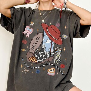 May include: A charcoal grey t-shirt featuring a graphic print of a cow-print cowboy boot, a red hat, a bow, and blocks with the letters T, S, and the number 13. The design includes the text TAYLOR and I KNEW YOU WERE TROUBLE on a beaded string.