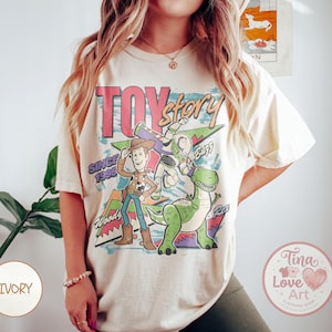 Puede incluir: Camiseta de color marfil con un gráfico de Woody, Buzz Lightyear y Rex de Toy Story. El diseño incluye el texto "Toy Story Since 1995" en un estilo retro. La camiseta tiene un corte holgado.