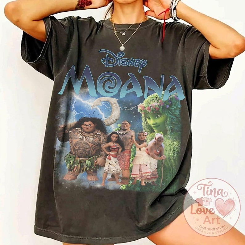 May include: Dark gray t-shirt featuring the Disney movie "Moana." The design includes the movie title in blue and the characters Moana, Maui, and others. The shirt has a vintage, worn look.