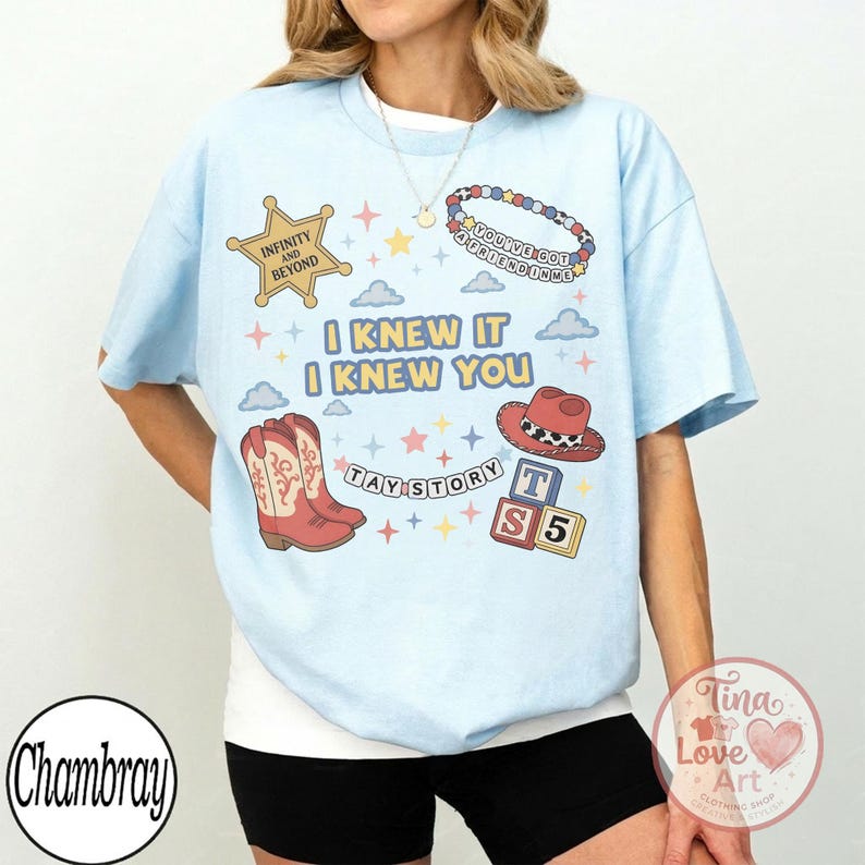 May include: A light blue t-shirt featuring a graphic print with the text I KNEW IT I KNEW YOU, TAY STORY, INFINITY AND BEYOND, and YOU'VE GOT A FRIEND IN ME. The design includes a cowboy boot, a cowboy hat, alphabet blocks, and colorful stars.