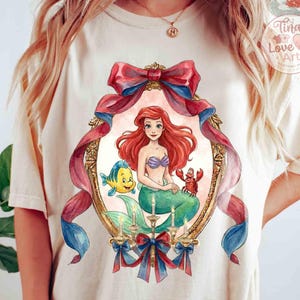 May include: Cream-colored t-shirt featuring a watercolor illustration of Ariel from The Little Mermaid. The artwork includes Ariel, Flounder, and Sebastian within an ornate frame with red and blue ribbons. The design is from Tina Love Art.