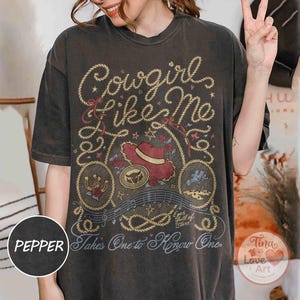 Pu&ograve; includere: Una t-shirt grigio antracite a maniche corte con una stampa grafica che riporta il testo Cowgirl like Me e Takes One to Know One. Il design include un cappello da cowboy rosso, un ferro di cavallo, un teschio di toro e bordi a corda.