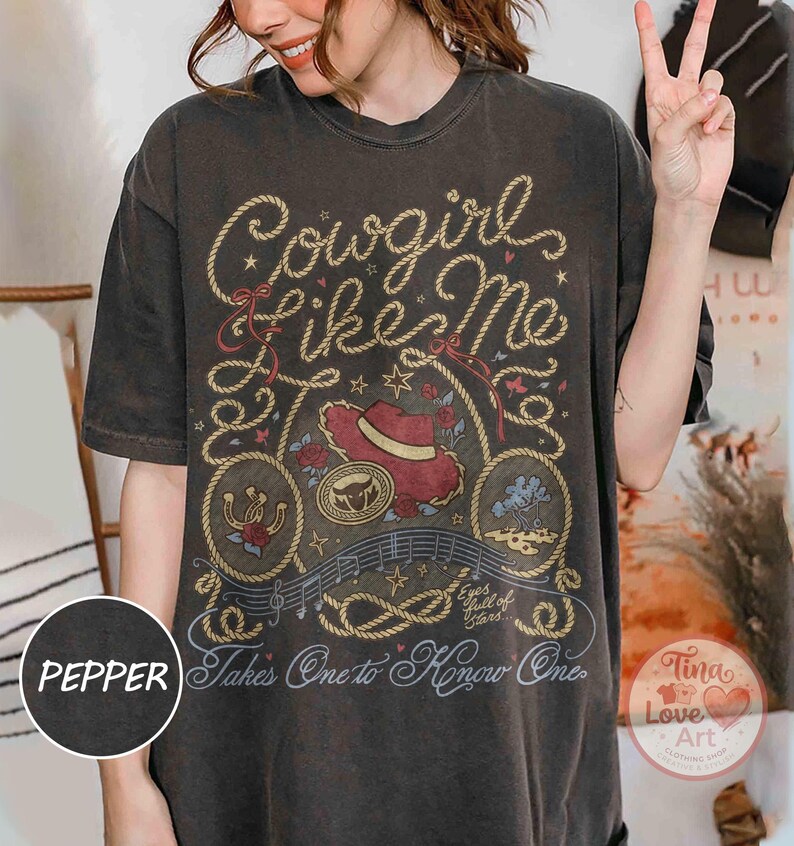 Pu&ograve; includere: Una t-shirt grigio antracite a maniche corte con una stampa grafica che riporta il testo Cowgirl like Me e Takes One to Know One. Il design include un cappello da cowboy rosso, un ferro di cavallo, un teschio di toro e bordi a corda.