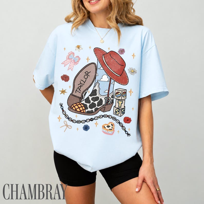 May include: A light blue t-shirt featuring a graphic print of a cow-print cowboy boot, a red hat, building blocks with the letters T, S, and 13, a pink bow, and a beaded string. The word TAYLOR is printed on the boot sole in a distressed style.