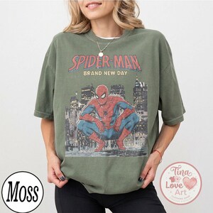 May include: Olive green t-shirt featuring a graphic of Spider-Man in a crouching pose against a cityscape backdrop. The text "SPIDER-MAN BRAND NEW DAY" is printed above the image in red and white. The shirt has short sleeves.