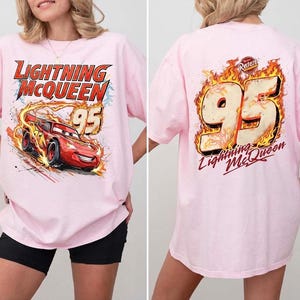 May include: A light pink t-shirt featuring a graphic of the red race car Lightning McQueen with the text Lightning McQueen and 95 on the front, and a large 95 graphic surrounded by flames with the text Lightning McQueen on the back.