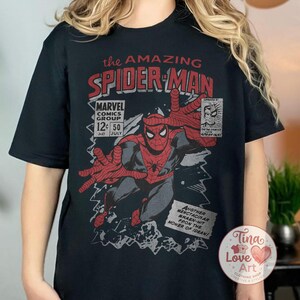Może przedstawiać: Czarna koszulka z nadrukiem w stylu vintage z komiksu Spider-Man. Grafika zawiera tekst "the AMAZING SPIDER-MAN" i "MARVEL COMICS GROUP". Projekt jest w kolorach czerwonym, czarnym i szarym.