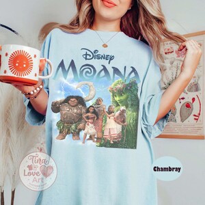 May include: Light blue t-shirt featuring the Disney movie "Moana" with a graphic of the characters. The shirt has the word "Disney" above "Moana" in a stylized font. The shirt is labeled "Chambray".