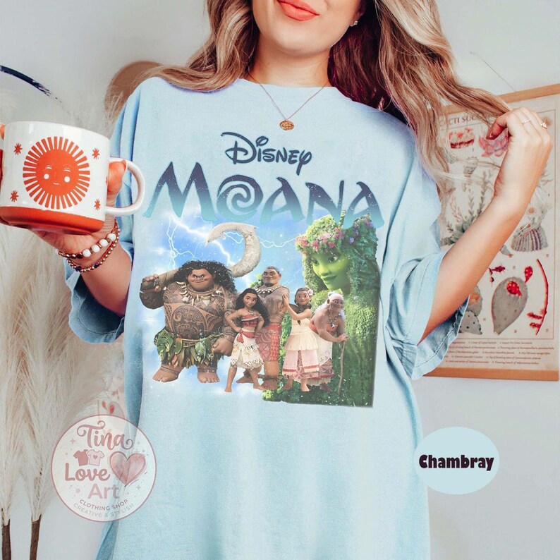 May include: Light blue t-shirt featuring the Disney movie "Moana" with a graphic of the characters. The shirt has the word "Disney" above "Moana" in a stylized font. The shirt is labeled "Chambray".