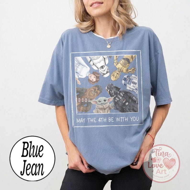 May include: Blue jean t-shirt featuring a Star Wars graphic. The graphic includes characters like Darth Vader, Yoda, and R2-D2, with the text "MAY THE 4TH BE WITH YOU" in a white frame. The shirt is a light blue color.