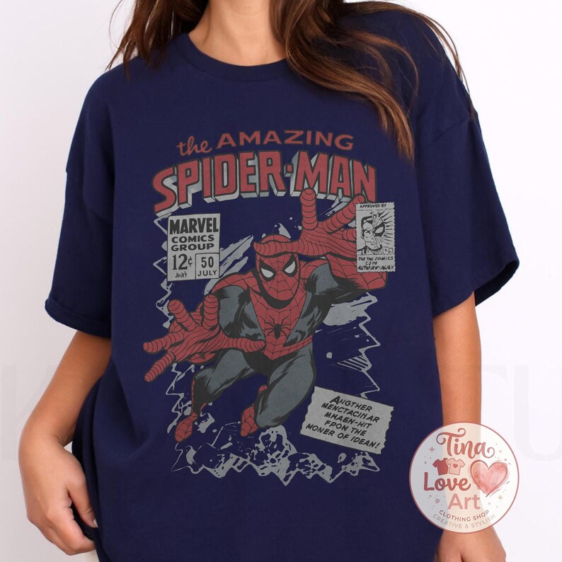 Może przedstawiać: Granatowa koszulka z nadrukiem w stylu vintage z komiksu Spider-Man. Grafika zawiera tekst "the AMAZING SPIDER-MAN" i "MARVEL COMICS GROUP". Obraz przedstawia Spider-Mana wyciągającego ręce.