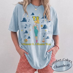May include: A light blue t-shirt featuring a graphic print of a blonde character in a teal leotard, surrounded by musical notes, clouds, a yellow star, a ball, and a traffic cone. The text TS and I Knew It, I Knew You are printed in yellow.