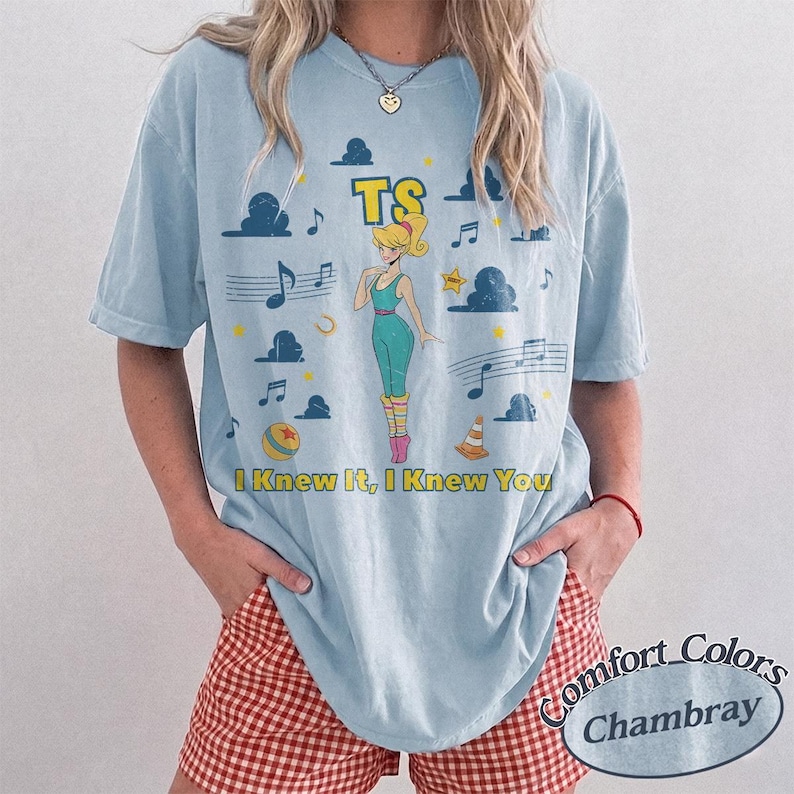 May include: A light blue t-shirt featuring a graphic print of a blonde character in a teal leotard, surrounded by musical notes, clouds, a yellow star, a ball, and a traffic cone. The text TS and I Knew It, I Knew You are printed in yellow.