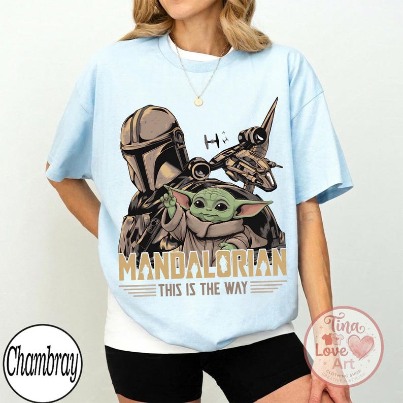 Pu&ograve; includere: T-shirt azzurra con grafica di The Mandalorian e Grogu, un'astronave e la scritta "MANDALORIAN THIS IS THE WAY". Il design utilizza tonalit&agrave; marroni, verdi e grigie.