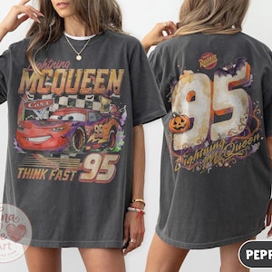 May include: A charcoal gray t-shirt featuring a graphic of Lightning McQueen from Cars on the front with the text Lightning McQueen and Think Fast 95. The back displays a large number 95 with a pumpkin, a bat, and the text Lightning McQueen.