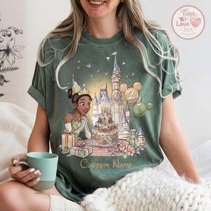 May include: Green t-shirt with a cartoon illustration of a princess, a castle, a birthday cake, and balloons. The number 2 is on the balloons. The text "Custom Name" is printed on the shirt.