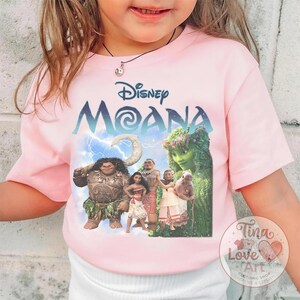 May include: Pink t-shirt featuring the Disney movie "Moana." The shirt displays the movie title in blue and white, along with a graphic of the characters. The shirt has short sleeves and a crew neck.