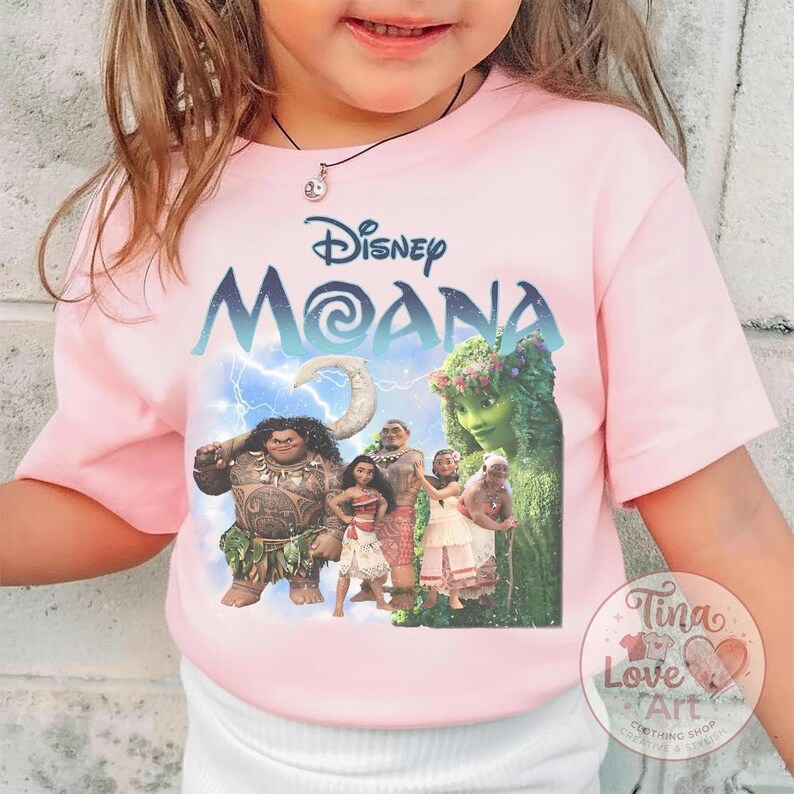 May include: Pink t-shirt featuring the Disney movie "Moana." The shirt displays the movie title in blue and white, along with a graphic of the characters. The shirt has short sleeves and a crew neck.