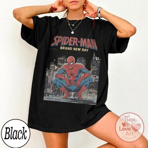 May include: Black t-shirt featuring a graphic of Spider-Man in a crouching pose, with the text "SPIDER-MAN BRAND NEW DAY" in red and blue. The design includes a cityscape backdrop. The shirt also has a round black tag with the word "Black".