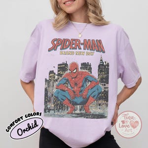 May include: Orchid-colored t-shirt featuring a graphic of Spider-Man in a crouching pose against a city skyline. The text "SPIDER-MAN BRAND NEW DAY" is above the image. The shirt has a vintage, distressed look.