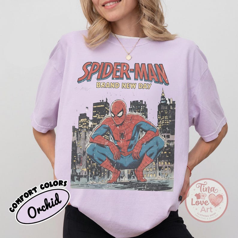 May include: Orchid-colored t-shirt featuring a graphic of Spider-Man in a crouching pose against a city skyline. The text "SPIDER-MAN BRAND NEW DAY" is above the image. The shirt has a vintage, distressed look.