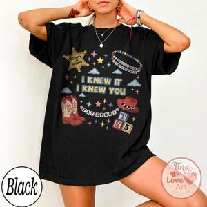 May include: A black short-sleeve t-shirt featuring a graphic print with the text I KNEW IT I KNEW YOU, TAY STORY, INFINITY AND BEYOND, and YOU'VE GOT A FRIEND IN ME. The design includes a sheriff badge, cowboy boots, a hat, and alphabet blocks.