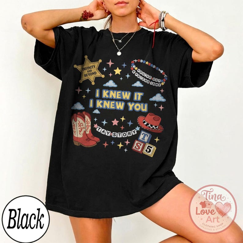 May include: A black short-sleeve t-shirt featuring a graphic print with the text I KNEW IT I KNEW YOU, TAY STORY, INFINITY AND BEYOND, and YOU'VE GOT A FRIEND IN ME. The design includes a sheriff badge, cowboy boots, a hat, and alphabet blocks.