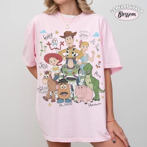 May include: A light pink t-shirt featuring a graphic print of Toy Story characters including Woody, Buzz Lightyear, Jessie, Rex, Hamm, Mr. Potato Head, Forky, and Bo Peep, with their names written in cursive script and small yellow stars.
