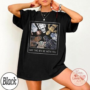 May include: Black t-shirt featuring a framed graphic of Star Wars characters including Darth Vader, Yoda, and R2-D2. The text "MAY THE 4TH BE WITH YOU" is printed below the graphic. The shirt has a relaxed fit.