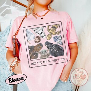May include: A light pink t-shirt featuring a framed graphic of Star Wars characters, including Darth Vader, Chewbacca, and Baby Yoda. The text "MAY THE 4TH BE WITH YOU" is printed below the image. The shirt is a casual style.