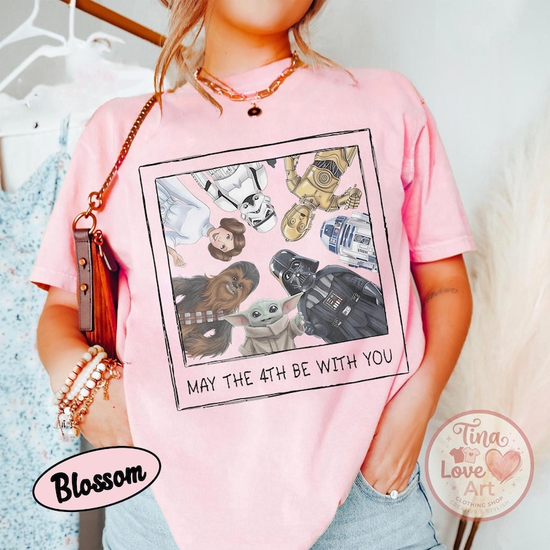 May include: A light pink t-shirt featuring a framed graphic of Star Wars characters, including Darth Vader, Chewbacca, and Baby Yoda. The text "MAY THE 4TH BE WITH YOU" is printed below the image. The shirt is a casual style.