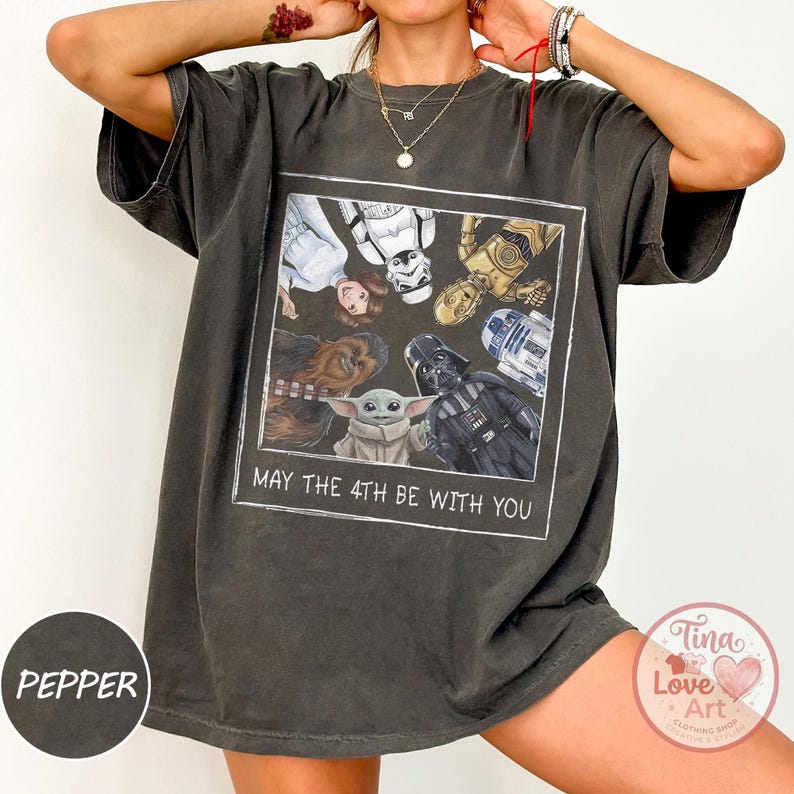 May include: Dark grey t-shirt featuring a Star Wars graphic. The graphic includes characters like Darth Vader, Baby Yoda, and R2-D2, with the text "MAY THE 4TH BE WITH YOU" in a white rectangle.
