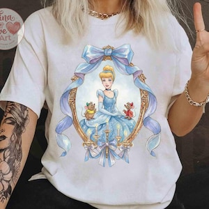 May include: White t-shirt featuring a framed illustration of Cinderella in a blue gown, with two mice. The frame is gold with a blue ribbon and bow. The shirt has the text "Tina Love Art Clothing Shop" in a heart-shaped logo.