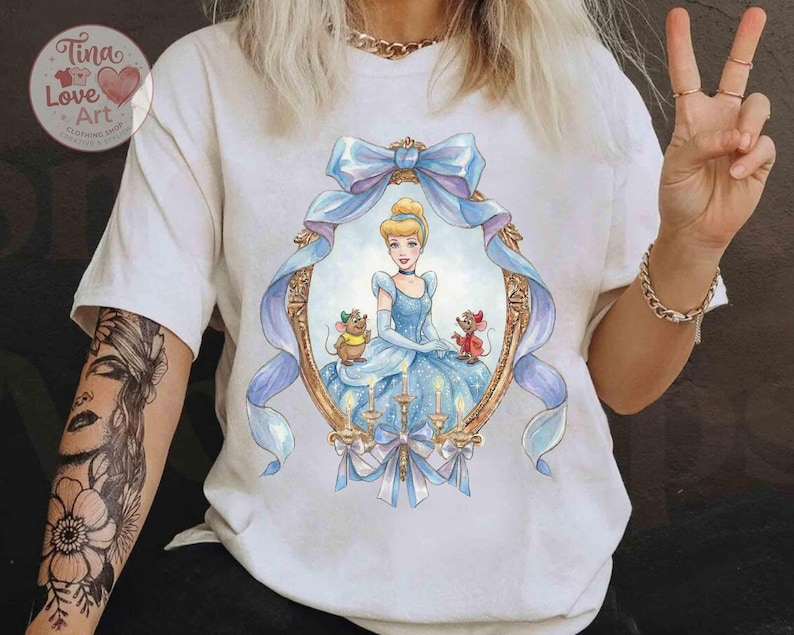 May include: White t-shirt featuring a framed illustration of Cinderella in a blue gown, with two mice. The frame is gold with a blue ribbon and bow. The shirt has the text "Tina Love Art Clothing Shop" in a heart-shaped logo.