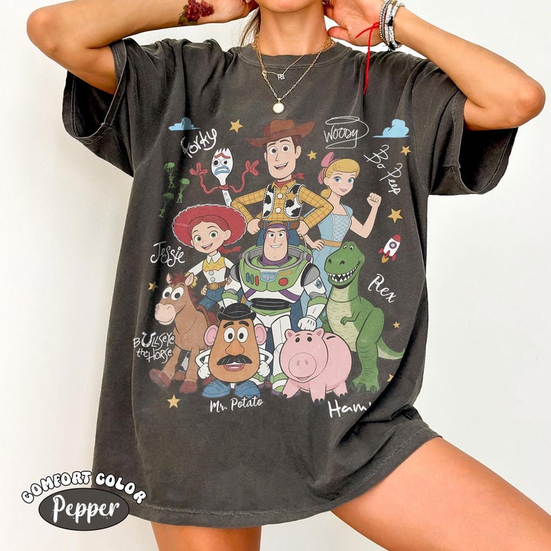 May include: A charcoal gray short-sleeve t-shirt featuring a graphic print of Toy Story characters including Woody, Buzz Lightyear, Jessie, Bullseye, Mr. Potato Head, Hamm, Bo Peep, and Forky, with their names written in a cursive script font.