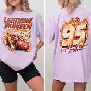 May include: A light purple t-shirt featuring a graphic of the red race car Lightning McQueen with the number 95 on the front. The back of the shirt displays the number 95 surrounded by flames and the text Lightning McQueen and Rust-eze.